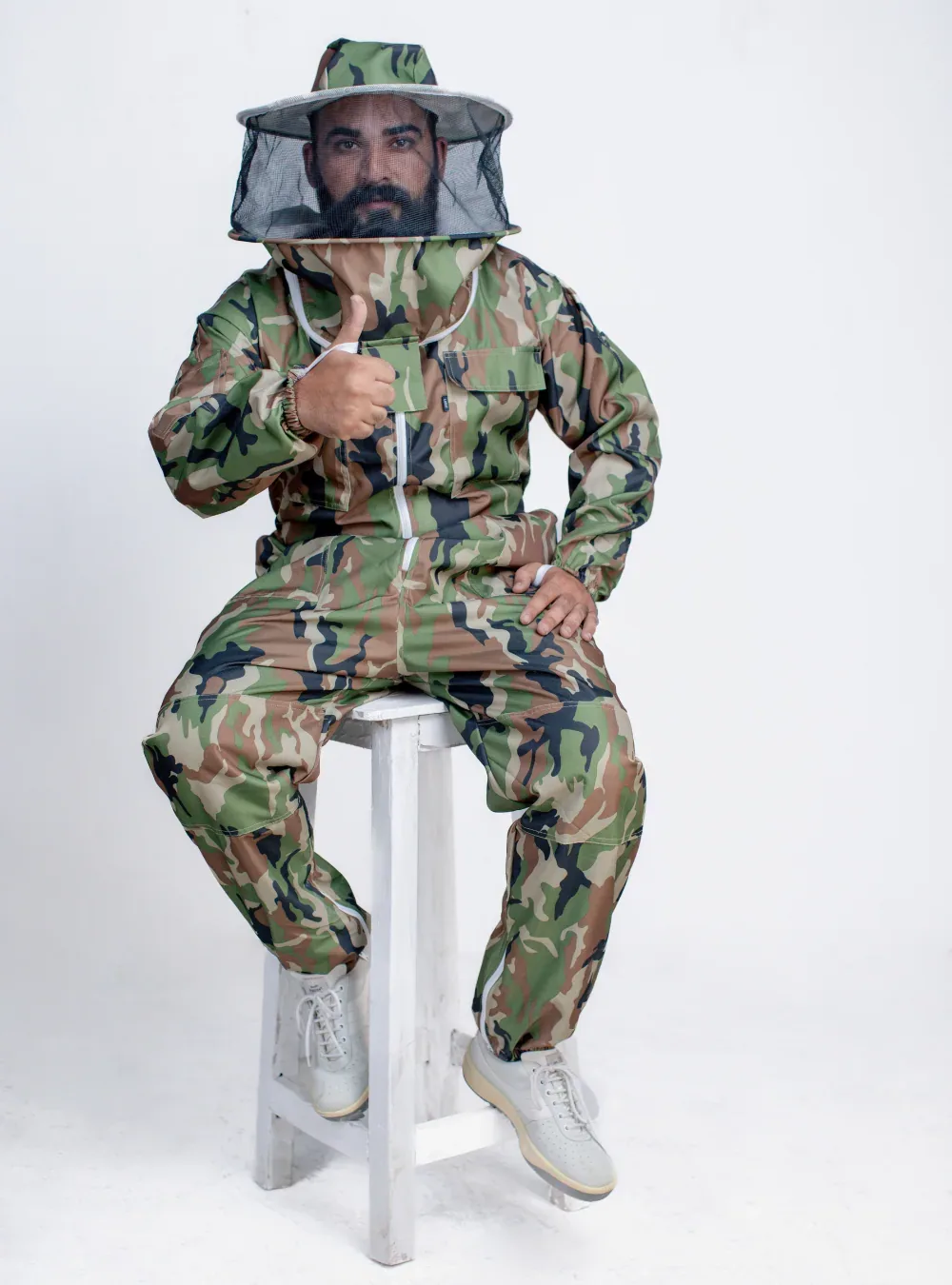 Beekeeper seated on a stool in a camouflage bee suit with a round veil, showing room at the knees and thighs